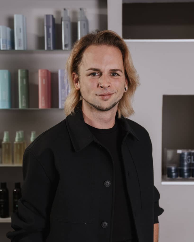 daniel farbexperte bei helena conzen hairdesign Daniel im Porträt: Blond- & Balayage-Profi bei deinem Friseur in Bochum Friseur Daniel im Salon von Helena Conzen Hairdesign in Bochum vor einem Regal mit Haarpflegeprodukten