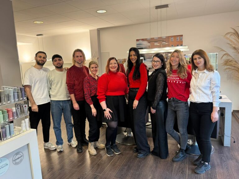 team moment im salon weihnachtszeit bei uns Wir lieben Haare Teammitglieder stehen lächelnd im Salonbereich und posieren gemeinsam für ein Foto; helle, moderne Einrichtung im Hintergrund.